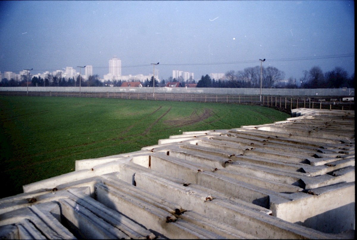 Berlin, 1990 – Auf dem Boden aneinandergereihte Mauerelemente vor dem Hinterlandzaun bei Großziethen (Fotograf: Albrecht Roos)