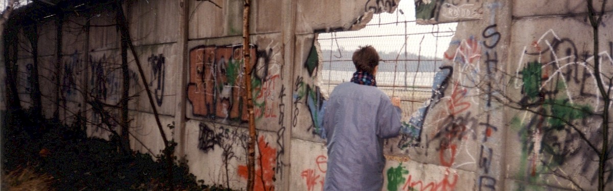 Berlin, Mitte Januar 1990 – Eine Frau blickt durch ein Loch in der Betonplattenmauer in Lichtenrade (Fotograf: Monika Waack)