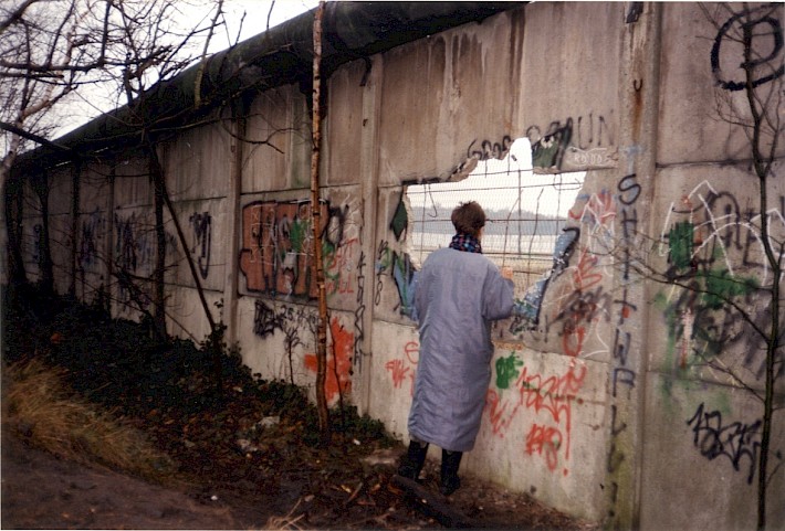 Berlin, Mitte Januar 1990 – Eine Frau blickt durch ein Loch in der Betonplattenmauer in Lichtenrade (Fotograf: Monika Waack)